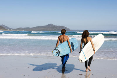 Rear view of friends carrying surfboards at beach during sunny day