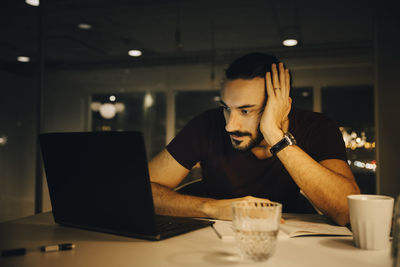 Tired businessman working late while staring at laptop on illuminated desk in office