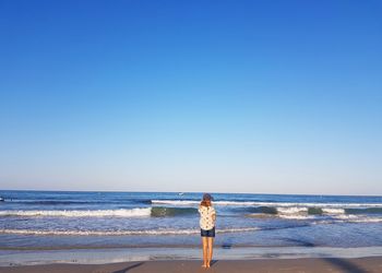 Rear view of girl standing at beach against clear blue sky