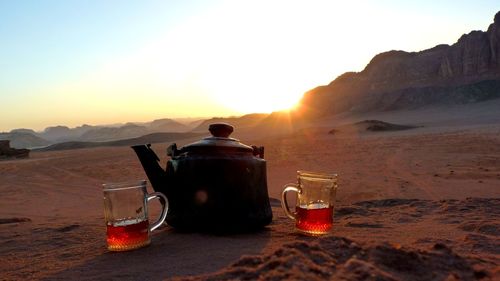 Tea kettle and glasses on desert against sky during sunset