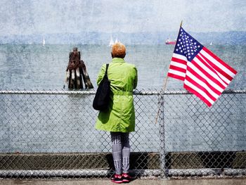 Rear view of woman standing by american flag on fence at san francisco bay