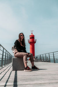 Young woman sitting on railing by sea against sky