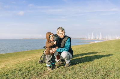 Grandfather and granddaughter looking away on land by sea against sky