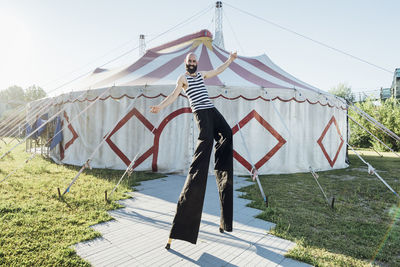 Male artist with arms outstretched walking with stilts on footpath