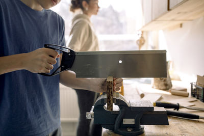 Midsection of male teenage student cutting wood with saw in carpentry class