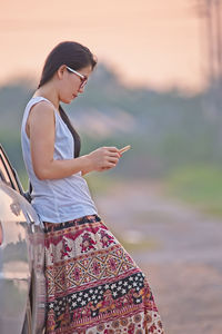 Side view of woman using phone while standing by car during sunset