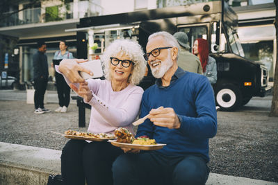 Smiling senior woman taking selfie with man eating meal against food truck in city