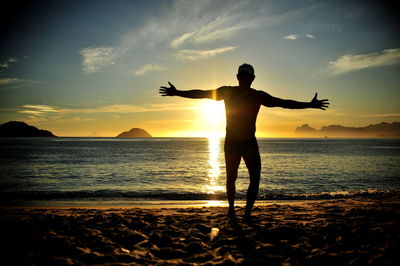 Silhouette man with arms outstretched standing at beach against sky during sunset