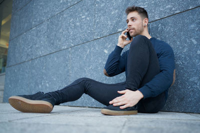 Side view of modern male in casual wear sitting near stone wall and br