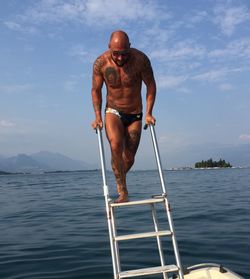 Full length of shirtless man standing by sea on ladder