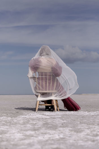 Rear view of teenage girl with scarf sitting on chair at salt flat against sky