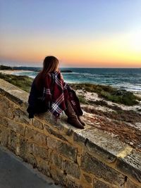 Full length of woman looking at sea while relaxing on retaining wall during sunset