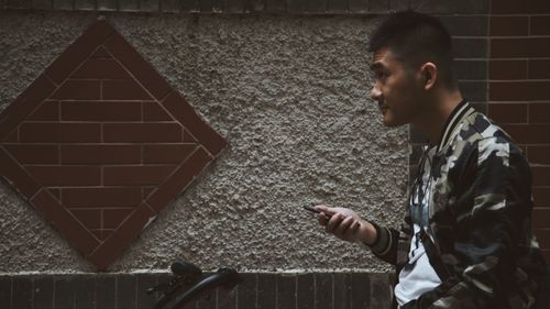 Side view of young man using mobile phone while sitting on bicycle against wall