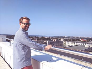 Side view of mid adult man wearing sunglasses while standing on building terrace against clear blue sky