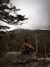 Full length of man sitting on rock by tree mountain against cloudy sky