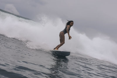 Side view of woman in wetsuit surfing on sea against cloudy sky