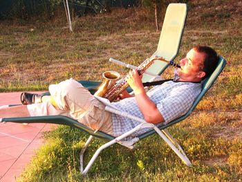 Side view of man playing saxophone while relaxing on lounge chair at grassy field