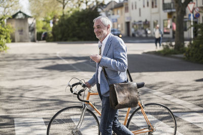 Smiling businessman with bicycle crossing road on sunny day