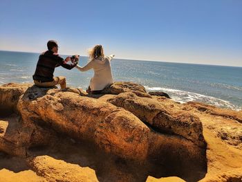 Couple on sandstone boulders at ocean whale watching woman pointing out to sea.