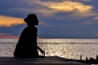 Silhouette woman sitting by sea against sky