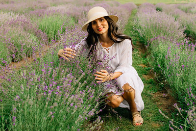 Close up of beautiful smiling woman in hat sitting on grass in lavender field 