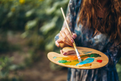 Midsection of woman holding paintbrush and palette while standing outdoors