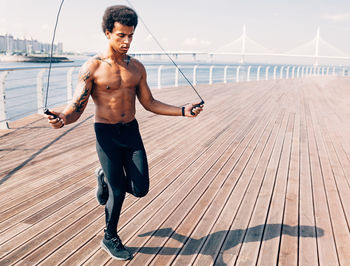 Full length of shirtless young man jumping rope on promenade
