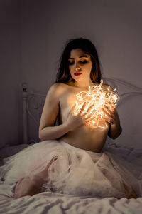 Sensuous young woman covering breasts with illuminated string lights on bed at home