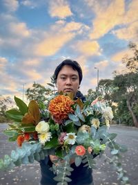 Portrait of young asian man with bouquet of fresh flowers and leaves against cloudy blue sky.