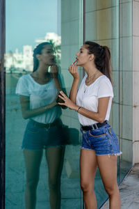 Beautiful woman applying lipstick while standing against glass wall