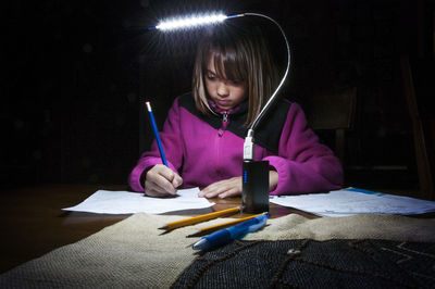 Girl studying by electric lamp on table in darkroom at home
