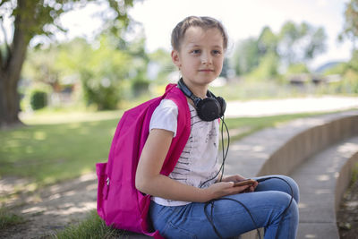 A cute happy girl with a pink backpack sits in a schoolyard  with headphones around her neck