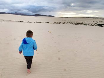 Rear view of boy walking at sandy beach against sky