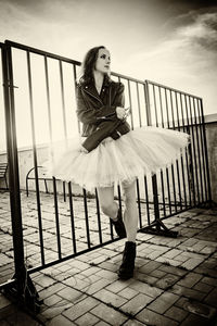 A ballerina in tutu stands at the fence on the roof at sunset in boots and a jacket