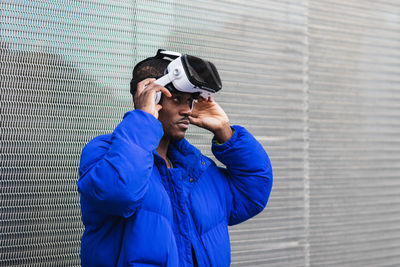 African american male standing on street near metal fence while exploring virtual reality in innovative goggles and touching air