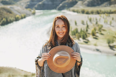 Carefree brunette young woman traveler in poncho with felt hat on background of mountain river