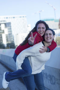 Portrait of smiling woman carrying friend while gesturing peace sign in city