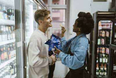 Side view of girlfriend feeding chips to boyfriend while standing at aisle at supermarket