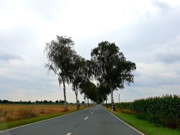 Empty road amidst trees on field against sky