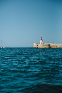 Lighthouse by sea against clear blue sky