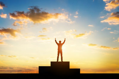 Silhouette man with arms raised standing winners podium against sky during sunset