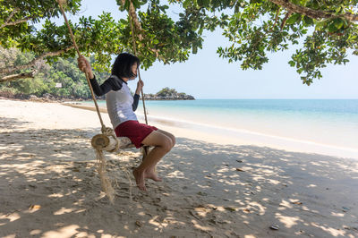 Side view of young woman swinging at beach against clear sky