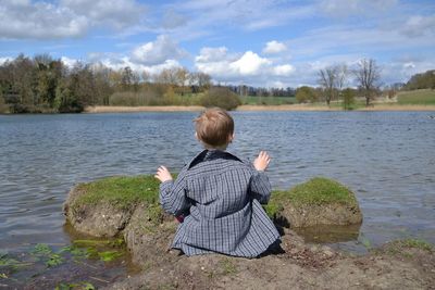 Rear view of boy sitting by lake against sky