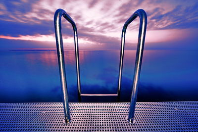 Close-up of metal ladder by swimming pool against sky during sunset