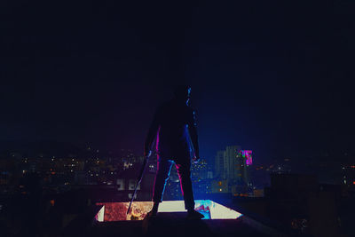 Rear view of man standing by illuminated buildings against sky at night