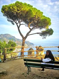 Woman sitting on bench by tree against sky