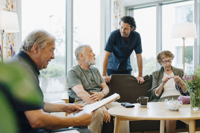 Male healthcare worker talking to senior man sitting on sofa amidst friends at retirement home