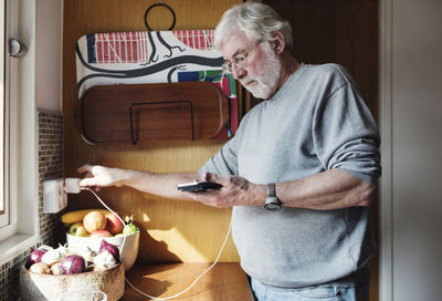 Senior man holding mobile phone while plugging charger to electrical outlet at domestic kitchen