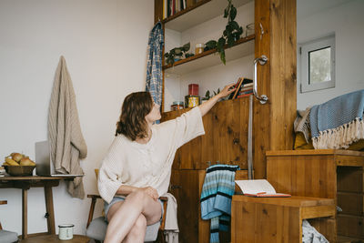 Young woman choosing book from rack while sitting on chair at home