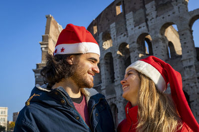 Roman holidays at christmas. a young couple poses in front of the colosseum in red santa claus hats.